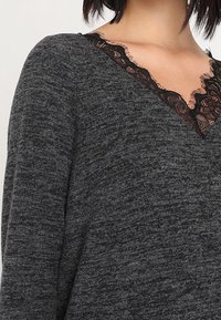 Pull en maille grise avec un col en V, orné d'une bordure en dentelle noire au niveau de l'encolure. Texture douce et coupe décontractée.