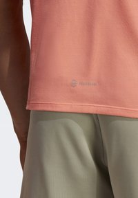 Camicetta color pesca a maniche corte con una trama liscia, caratterizzata da un logo Aeroready vicino all'orlo; abbinata a pantaloncini sportivi verde chiaro.