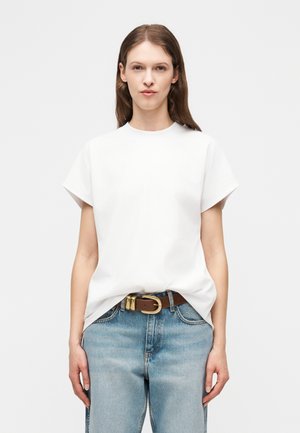 Νεαρή γυναίκα με ίσια καστανά μαλλιά φοράει άσπρο χαλαρό μπλουζάκι με κοντά μανίκια που είναι μαζεμένο μέσα σε ανοιχτόχρωμο τζιν και καφέ ζώνη.