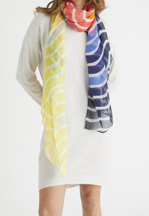 Femme portant une robe pull beige clair et une longue écharpe à rayures ondulées jaune, orange, bleue et bleu marine drapée autour du cou.