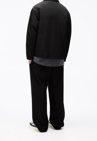 Czarna kurtka typu bomber, szara warstwowa koszula, luźne czarne spodnie oraz czarno-białe trampki, widoczne od tyłu na jednolitym białym tle.