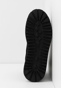 Semelle de chaussure noire avec des crampons profonds et des motifs en forme de croix, se tenant debout contre un mur d'angle blanc et un sol blanc.