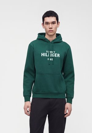 Jeune homme portant un sweat à capuche vert foncé avec un texte blanc "Tommy Hilfiger 1985", debout devant un fond gris clair uni.