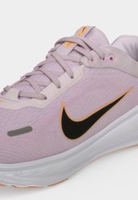 Ανοιχτό μοβ παπούτσι για τρέξιμο Nike με μαύρο και πορτοκαλί λογότυπο swoosh, λευκή σόλα και κορδόνια σε λευκό φόντο.