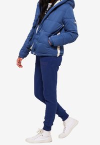 Blauwe pufferjack met capuchon, witte accenten, gecombineerd met donkerblauwe trainingsbroek en witte sneakers. Glad materiaal en een ontspannen pasvorm.