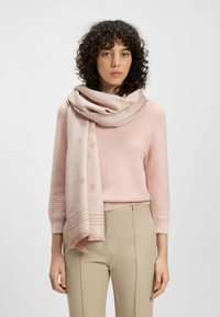 Jasnoróżowy szalik wykonany z miękkiego materiału, z subtelnymi wzorami i frędzlami, ułożony na szyi modelki. Uzupełnia swobodny strój.