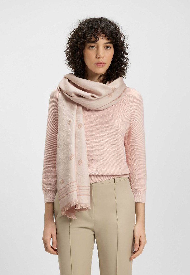 Jasnoróżowy szalik wykonany z miękkiego materiału, z subtelnymi wzorami i frędzlami, ułożony na szyi modelki. Uzupełnia swobodny strój.