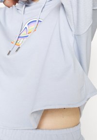 Hoodie accorciato blu chiaro con cordini e punte metalliche. Presenta un design grafico multicolore sulla parte frontale. Texture morbida.