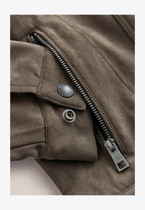 Pruun seemisnahast jakk, millel on hõbedane lukk, mustad nööbid ja kuldsed aktsendid. Pehme tekstuur, üksikasjaliku õmblusega ja sile disain.