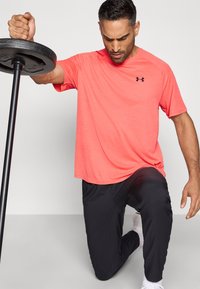 Kirkas korallinvärinen treenipaita, jossa on lyhyet hihat ja hienovarainen tekstuuri, yhdistettynä mustiin kevyisiin housuihin. Nostamassa painolevyä.