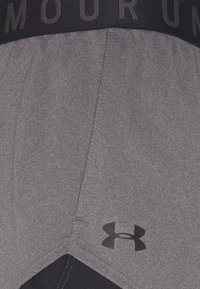 Gros plan sur un short de sport gris avec un logo Under Armour noir près de l'ourlet et une ceinture noire avec un texte partiel dessus.