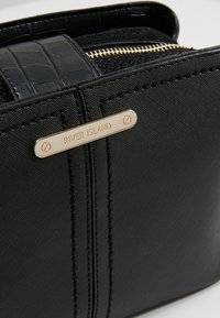 Svart handväska i texturerat tyg med krokodilsmönster, guldfärgad hårdvara och en liten rektangulär etikett som säger "RIVER ISLAND."