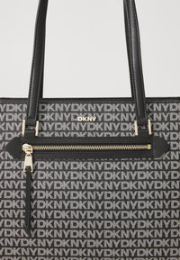 DKNY tote bag with black and white logo pattern, featuring a top zip closure, gold-tone hardware, and two sturdy handles.