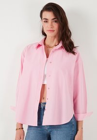 Pink button-up shirt with a collared neckline, long sleeves, and a relaxed fit. Made from a smooth fabric, featuring a slight sheen.