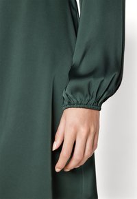 Pitkähihaista mekkoa tummanvihreästä kankaasta, jossa on sileä pinta. Hihoissa on kokoontuva nielu ja käsi lepää kankaan päällä.