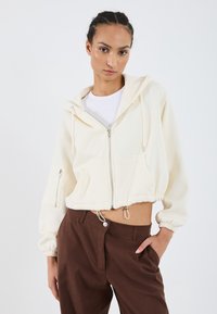 Korte crème hoodie met een rits aan de voorkant, een trekkoord aan de onderkant en buidelzakken. G gedragen met bruine broek, die een gladde textuur en een ontspannen pasvorm heeft.