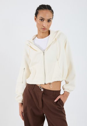 Kurze, cremefarbene Kapuzenjacke mit Reißverschluss, Kordelzugsaum und Kängurutaschen. Getragen mit braunen Hosen, die eine glatte Textur und eine lockere Passform aufweisen.