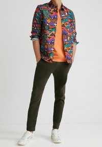 Camicia a fantasia multicolore in viola, arancione e teal sopra una t-shirt arancione, abbinata a pantaloni verde scuro e sneakers bianche con disegni.