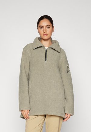 Pull en polaire de couleur vert olive clair, avec un col zippé, des manches longues et une patch d'accent sur le côté du bras gauche. Texture douce.