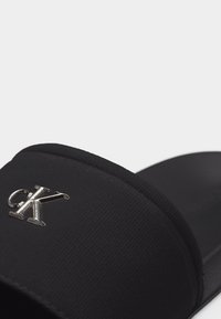 Sandale à glissière noire avec bride en tissu texturé présentant un logo "ck" métallique en relief sur la partie supérieure.