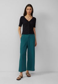 Zwarte kortom cardigan met een golvende halslijn en knopen, gecombineerd met teal wijde broekspijpen en zwarte gespsandalen.