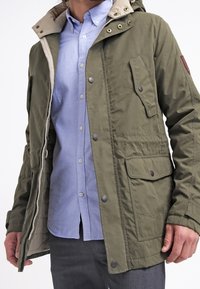 Olive green, water-resistant jacket with a beige hood. Features zip closure, chest pocket, and two front pockets. Lightweight fabric, relaxed fit.