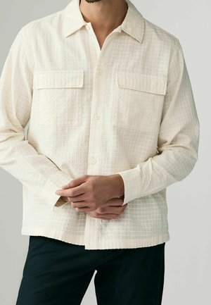 Homme portant une chemise crème texturée à boutons avec deux poches poitrine et un pantalon foncé, les mains légèrement jointes à la taille.