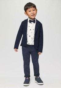 Marineblazer over een witte shirt met kleine zwarte patronen, gecombineerd met marineblauwe broek en grijze sneakers met rode accenten. Vlinderdas inbegrepen.