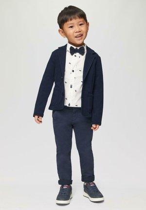 Marineblazer over een witte shirt met kleine zwarte patronen, gecombineerd met marineblauwe broek en grijze sneakers met rode accenten. Vlinderdas inbegrepen.