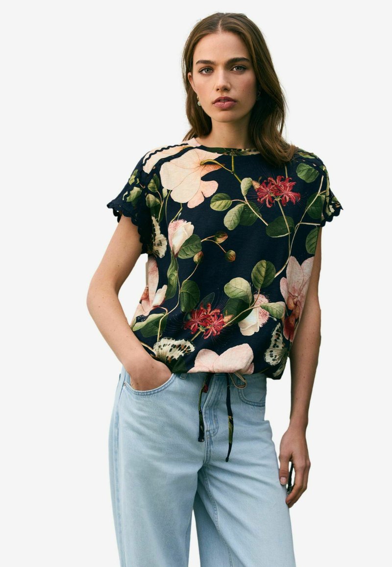Marinblå topp med blommönster och vågiga ärmar. Har ljusa blommor och gröna blad. Matchas med ljusblå vida jeans.