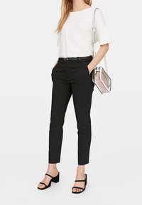 Zwarte tailormade broek met een slim fit, gecombineerd met een wit T-shirt. Accessoires zijn een doorzichtige handtas en zwarte sandalen met bandjes.