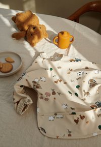 Beige children's bib with colorful farm-themed illustrations, made of waterproof material. Includes a pocket and elasticized cuffs.
