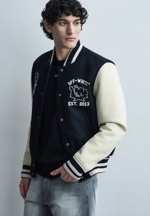 Νέος άνδρας με σγουρά μαλλιά που φοράει ένα μαύρο και λευκό varsity μπουφάν με την επιγραφή «Off-White EST. 2013» και ξεθωριαμένο γκρι τζιν, κοιτάζοντας πλάγια.