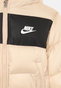 Μπεζ φουσκωτό μπουφάν με μαύρη γωνιακή επένδυση στο στήθος, μπροστινό φερμουάρ, πλαϊκή τσέπη, και λευκό λογότυπο Nike με σήμα στο στήθος.