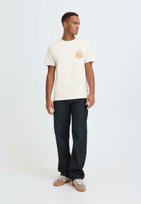 Roomkleurig katoenen T-shirt met een oranje "Fresh Blend"-opdruk op de linkerborst, gecombineerd met donkerblauwe spijkerbroek en grijze sneakers.