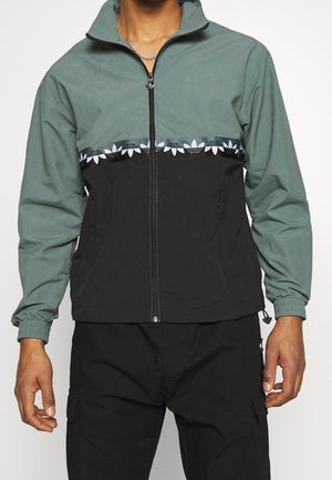Mann trägt eine grün-schwarze Zip-up-Jacke mit einem gemusterten Streifen über der Brust und schwarze Cargohosen.