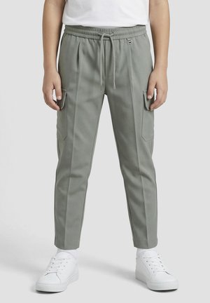 Enfant portant un pantalon cargo gris fuselé avec des poches latérales, une taille élastique avec cordon, des chaussettes blanches et des baskets blanches, debout sur un fond gris.