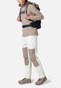 Beige softshell jakke med hætte, hvide og brune panelbukser og grå vandresko. Rygsækken har sorte net detaljer og spænder.