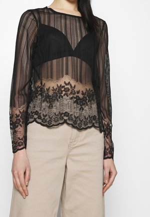 Vrouw draagt een zwarte doorschijnende top met lange mouwen en bloemmotieven in kant over een zwarte beha, gecombineerd met beige hoge taille broek.