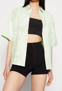 Camisa verde claro de manga corta con un patrón tie-dye, diseño abotonado, y un top corto texturizado negro combinado con pantalones cortos negros de tiro alto.