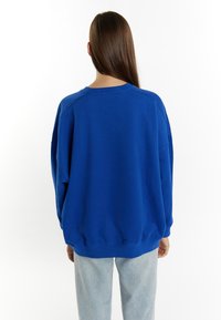 Felpa blu oversize con colletto e polsini a coste, caratterizzata da una texture morbida. Abbinata a pantaloni in denim azzurro.