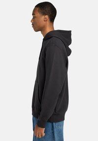 Schwarzer Kapuzenpullover mit Kängurutasche, gerippten Bündchen und Saum; getragen mit blauen Jeans, von der Seite betrachtet.