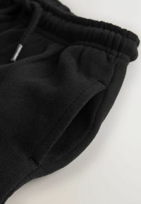 Črne bombažne trenirke z elastičnim pasom, stranskimi žepi in mehko teksturo. Prikaz v bližnjem načinu poudarja podrobnosti tkanine in šive.