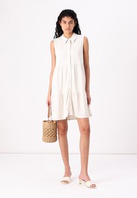 Robe sans manches crème avec fermeture à boutons sur le devant, taille froncée et tissu texturé ; associée à un panier tissé et des sandales de couleur claire.