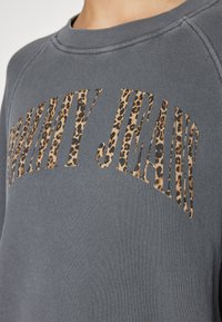 Grå sweatshirt med rundad halsringning har fet text "Vanity Jeans" i leopardmönster, vilket betonar en trendig, avslappnad stil.