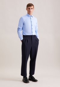 Camicia button-up a maniche lunghe color azzurro chiaro abbinata a pantaloni plissettati blu navy scuri e scarpe da uomo nere. Design semplice e su misura.