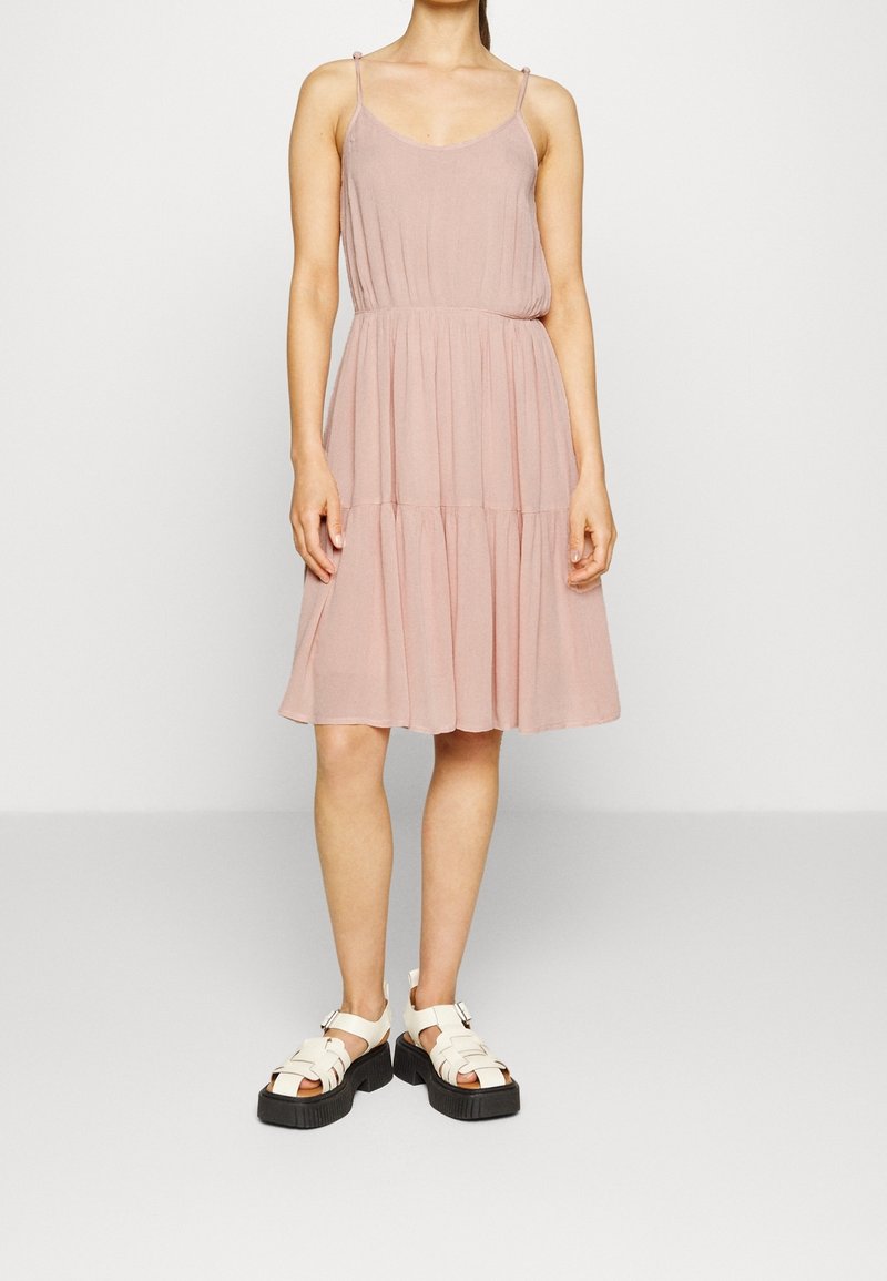 Femme portant une robe sans manches rose clair, longueur genoux, associée à des sandales blanches épaisses sur un fond uni.