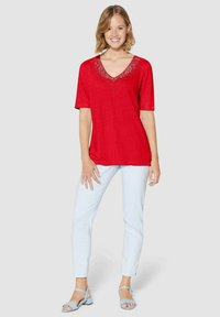 Rode blouse met korte mouwen en kantdetail bij de V-hals, gecombineerd met lichtblauwe gestreepte broek en blauwe sandalen, gepresenteerd tegen een neutrale achtergrond.