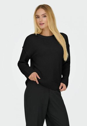 Maglione nero oversize con scollatura e orlo a costine, realizzato in tessuto morbido. Indossato con pantaloni neri larghi. Design semplice e casual.