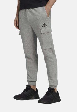 Uomo che indossa pantaloni della tuta cargo grigi Adidas con sneakers nere, in piedi con una mano in tasca su sfondo semplice.